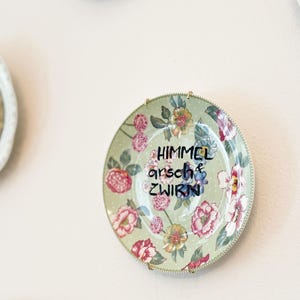 Könnte beinhalten: Ein runder dekorativer Keramikteller mit hellgrünem Hintergrund, weißen Punkten und einem Blumenmuster aus rosa, roten und gelben Blüten. In der Mitte des Tellers steht der schwarze Schriftzug HIMMEL arsch & ZWIRN.