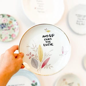 Könnte beinhalten: Ein weißer Keramikteller mit goldfarbenem Rand, verziert mit einer zarten botanischen Illustration von Blättern in Gelb-, Rosa- und Grautönen, mit dem schwarzen Text "AND UP GOES THE LUZIE" in der Mitte der Telleroberfläche gedruckt.