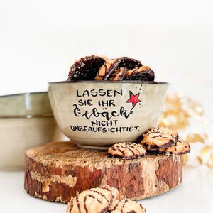 Könnte beinhalten: Eine Keramikschale gefüllt mit dunklen Keksen, mit dem Text "LASSEN SIE IHR GEBÄCK NICHT UNBEAUFSICHTIGT" und einem roten Stern. Die Schale steht auf einer Holzscheibe mit Keksen. Eine weitere Schale ist im Hintergrund.