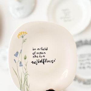 Könnte beinhalten: Ein weißer Keramikteller mit unregelmäßiger, organischer Form und einer handgemalten Illustration von gelben und blauen Wildblumen auf der linken Seite. Der Text "in a field of roses she is a wildflower" ist in schwarzer Schrift mittig platziert.