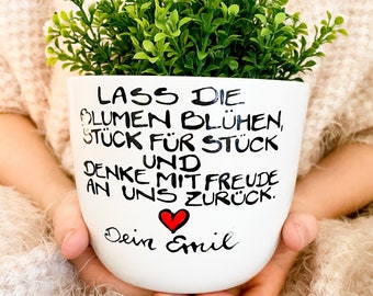 May include: White ceramic planter with black hand-painted lettering that reads "Lass die Blumen blühen, Stück für Stück und denke mit Freude an uns zurück. Dein Emil". A small red heart is painted above the name Emil. The planter is filled with a green plant and is being held by a person's hands.