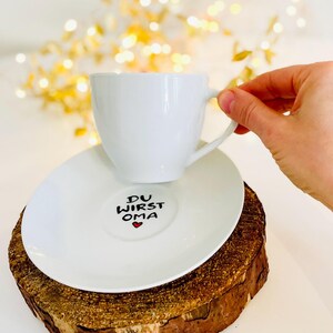 May include: A white teacup and saucer set with the text "DU WIRST OMA" and a red heart on the saucer. The cup is held by a hand. The set rests on a wooden slice, with a blurred background of golden lights.