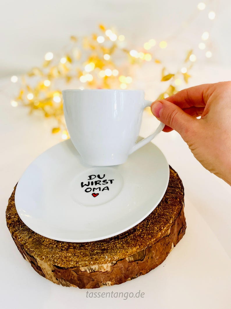 May include: A white teacup and saucer set with the text "DU WIRST OMA" and a red heart on the saucer. The cup is held by a hand. The set rests on a wooden slice, with a blurred background of golden lights.