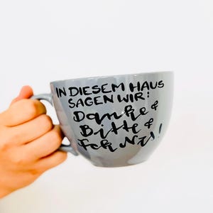 Könnte beinhalten: Eine graue Keramiktasse mit Henkel, die mit schwarzem, handgeschriebenem Text versehen ist: IN DIESEM HAUS SAGEN WIR: Danke & Bitte & Fck Nz!. Die Tasse hat eine glatte, abgerundete Form und wird von einer Hand gehalten, mit der URL tassentango.de unten.