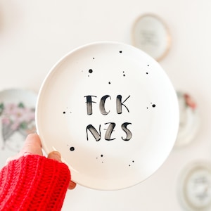 Könnte beinhalten: Weiße Keramikplatte mit schwarzem Text "FCK NZS" und verstreuten schwarzen Punkten. Die Platte wird von einer Person gehalten, die einen roten Pullover trägt. Weitere dekorative Teller sind im Hintergrund zu sehen.