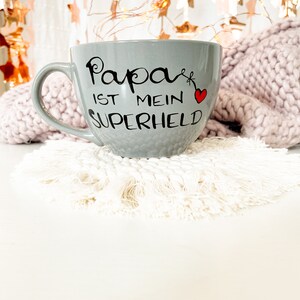 Op de afbeelding: Een grijze keramische mok met een zwarte inscriptie die "Papa ist mein Superheld" luidt met een rood hart. De mok staat op een witte gehaakte onderzetter.