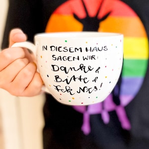 Könnte beinhalten: Eine weiße Keramiktasse mit bunten Punkten und dem handgeschriebenen schwarzen Text: IN DIESEM HAUS SAGEN WIR: Danke & Bitte & Fck Nts! Die Tasse hat einen geschwungenen Henkel und eine glatte, glänzende Oberfläche mit einer deutlichen Beschriftung.