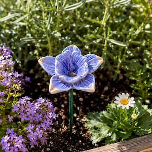 Könnte beinhalten: Ein Keramik-Gartenstecker in Form einer blauen, sechsblättrigen Blume mit goldfarbenen Rändern auf einem dünnen grünen Metallstab. Die Blume hat eine texturierte, gerippte Oberfläche und steckt in dunkler Erde zwischen verschiedenen Pflanzen.