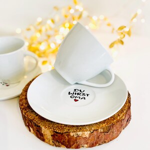 May include: White teacup and saucer set with the text "DU WIRST OMA" and a red heart on the saucer. The saucer rests on a rustic wooden coaster. A second teacup and saucer are visible in the background. The background has soft, blurred golden lights.