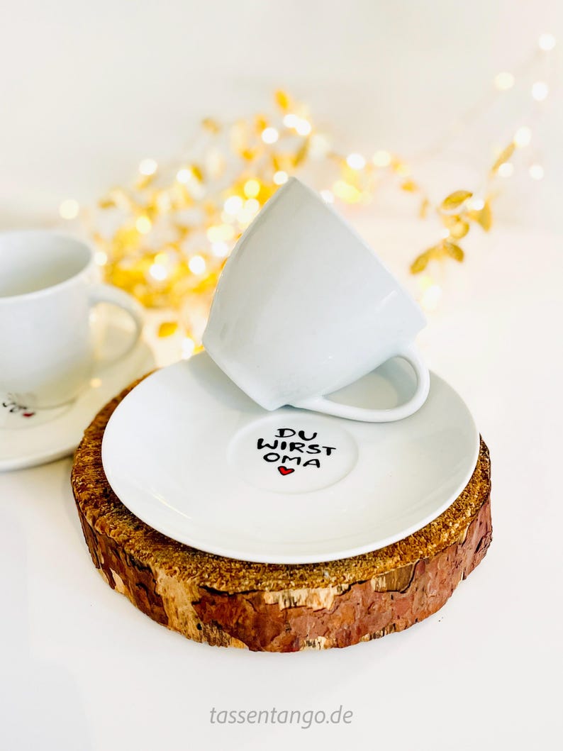May include: White teacup and saucer set with the text "DU WIRST OMA" and a red heart on the saucer. The saucer rests on a rustic wooden coaster. A second teacup and saucer are visible in the background. The background has soft, blurred golden lights.