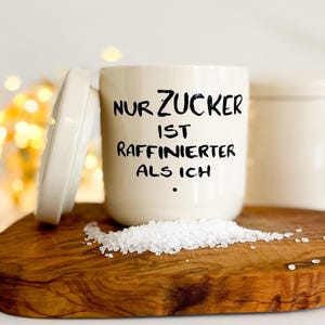 Könnte beinhalten: Eine cremefarbene Keramik-Zuckerdose mit passendem Deckel, bedruckt mit dem schwarzen Text NUR ZUCKER IST RAFFINIERTER ALS ICH. Vor der Dose liegt ein kleiner Haufen weißer Kristallzucker auf einer Holzoberfläche.