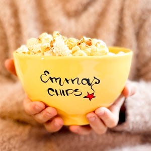 Könnte beinhalten: Eine gelbe Keramikschüssel mit den Worten "Emma's Chips" und einem roten Stern darauf. Die Schüssel ist mit Popcorn gefüllt.