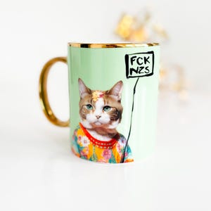 Könnte beinhalten: Ein hellgrüner Keramikbecher mit goldfarbenem Henkel und Rand. Der Becher zeigt die Grafik einer Katze in einem bunten Blumenhemd mit einer Blume auf dem Kopf, die ein schwarzes Schild mit dem weißen Text FCK NZS in einem Kasten hält.