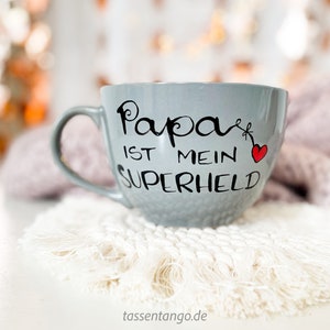 Op de afbeelding: Een grijze keramische mok met een zwarte inscriptie die "Papa ist mein Superheld" en een rood hartje zegt. De mok staat op een witte gehaakte onderzetter.