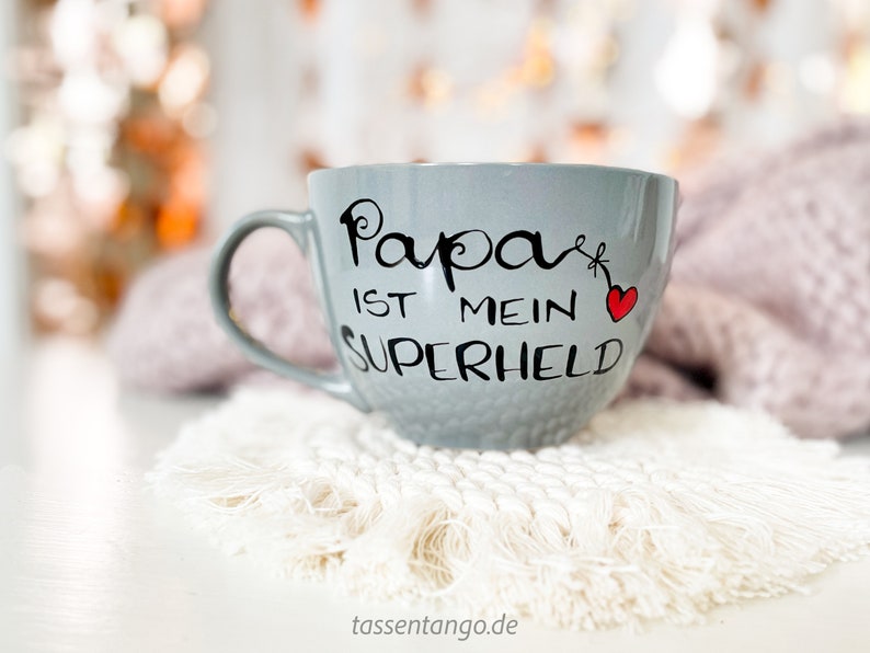 Op de afbeelding: Een grijze keramische mok met een zwarte inscriptie die "Papa ist mein Superheld" en een rood hartje zegt. De mok staat op een witte gehaakte onderzetter.
