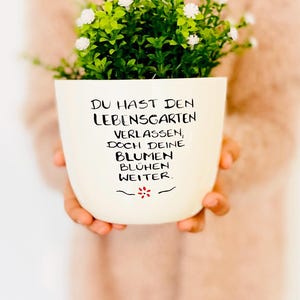 Könnte beinhalten: Ein cremefarbener Keramik-Blumentopf mit einer grünen Pflanze und weißen Blüten. Der Topf trägt den schwarzen Text "DU HAST DEN LEBENSGARTEN VERLASSEN, DOCH DEINE BLUMEN BLÜHEN WEITER." Unter dem Text ist eine rote Blume gezeichnet.