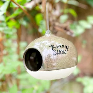 Könnte beinhalten: Ein keramisches Vogelhaus in Olivgrün mit weißem Sockel und dem Schriftzug "Piep Show" in Schwarz. Das runde Vogelhaus hat eine große Öffnung und hängt an einem dünnen Faden. Der Hintergrund ist ein verschwommenes Grün.