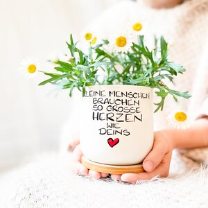 Könnte beinhalten: Eine weiße Keramiktasse mit einer handgeschriebenen Botschaft, die lautet "Kleine Menschen brauchen so grosse Herzen wie Deins" mit einem roten Herzen am Ende. Die Tasse ist mit weißen Gänseblümchen gefüllt und wird von einer Person gehalten.