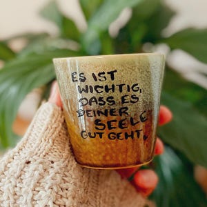 Könnte beinhalten: Ein Keramikbecher mit einer gesprenkelten braunen und beigen Glasur, der den handgemalten schwarzen deutschen Text trägt: ES IST WICHTIG, DASS ES DEINER SEELE GUT GEHT. Der Becher hat eine zylindrische Form mit einer strukturierten Oberfläche.