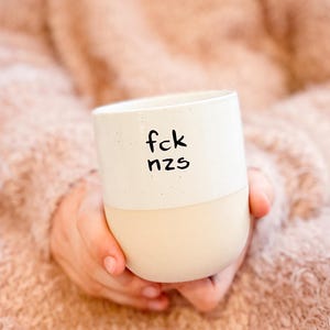Könnte beinhalten: Eine zweifarbige Keramiktasse mit cremefarbenem Boden und weißem Oberteil. Die Tasse trägt den Text "fck nzs" in Schwarz. Die Tasse wird in den Händen einer Person gehalten, mit einem weichen, strukturierten Hintergrund.