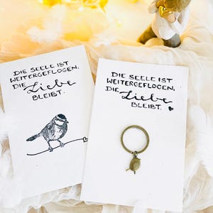 Könnte beinhalten: Zwei weiße Karten mit dem Text "DIE SEELE IST WEITERGEFLOGEN, DIE Liebe BLEIBT." Eine Karte zeigt eine Vogelillustration, die andere einen Vogel-Schlüsselanhänger. Der Hintergrund umfasst Lichterketten und eine Figur.