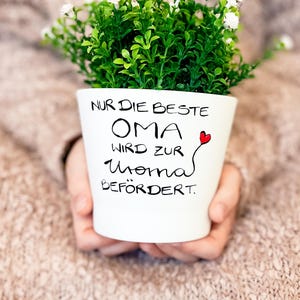 Könnte beinhalten: Weißer Keramik-Blumentopf mit dem Text "NUR DIE BESTE OMA WIRD ZUR MAMA BEFÖRDERT" in schwarzer Schrift und einem kleinen roten Herz. Der Topf ist mit grünem Laub und kleinen weißen Blüten gefüllt.