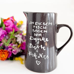 Könnte beinhalten: Ein dunkelgrauer Keramikkrug mit glänzender Oberfläche und einem Henkel. Auf der Vorderseite steht in weißer Handschrift: AN DIESEM TISCH SAGEN WIR: Danke & Bitte & Fck Nzs, gefolgt von einem kleinen Herzsymbol. Der Krug verfügt über einen Ausgießer.