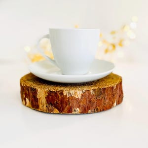 May include: A white teacup and saucer set atop a rustic, round wooden coaster. The coaster has a natural bark edge and a light brown color. The teacup is centered on the saucer, creating a simple, elegant presentation.