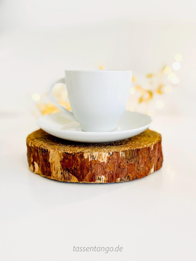 May include: A white teacup and saucer set atop a rustic, round wooden coaster. The coaster has a natural bark edge and a light brown color. The teacup is centered on the saucer, creating a simple, elegant presentation.