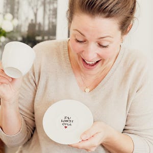 May include: A white teacup and saucer set. The saucer has the text "DU WIRST OMA" with a heart. The woman in the image is smiling and holding the teacup and saucer. The website tassentango.de is visible.
