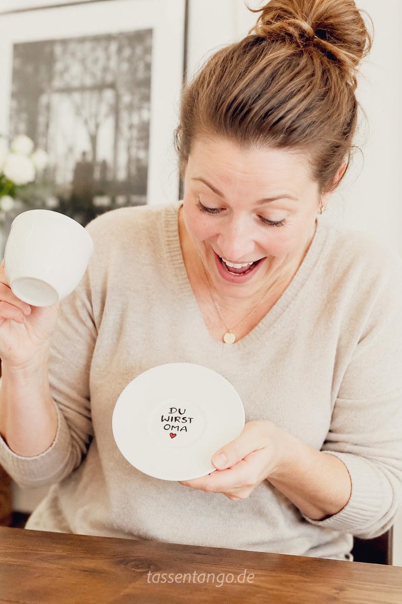 May include: A white teacup and saucer set. The saucer has the text "DU WIRST OMA" with a heart. The woman in the image is smiling and holding the teacup and saucer. The website tassentango.de is visible.