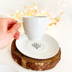 May include: A white teacup and saucer set with the text "DU WIRST OMA" and a red heart on the saucer. The cup is held by a hand. The set rests on a wooden coaster. The background has blurred golden lights.
