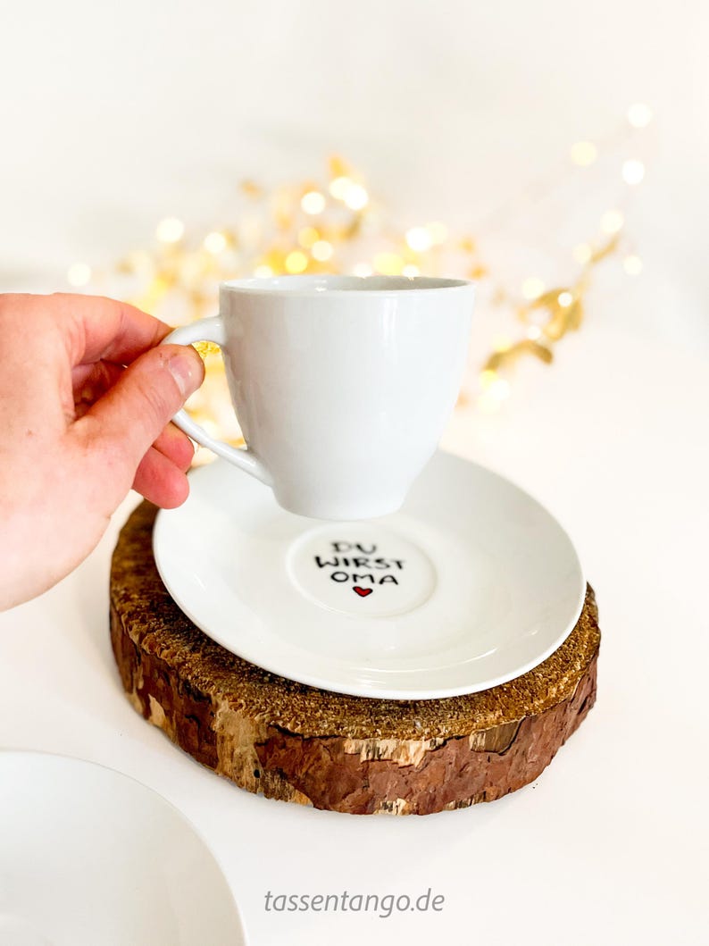 May include: A white teacup and saucer set with the text "DU WIRST OMA" and a red heart on the saucer. The cup is held by a hand. The set rests on a wooden coaster. The background has blurred golden lights.
