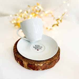 May include: A white ceramic teacup and saucer set with the text "DU WIRST OMA" and a heart symbol on the saucer. The set rests on a round, natural wood coaster. Decorative lights are in the background.