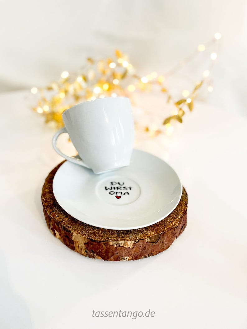 May include: A white ceramic teacup and saucer set with the text "DU WIRST OMA" and a heart symbol on the saucer. The set rests on a round, natural wood coaster. Decorative lights are in the background.