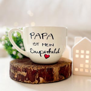 Op de afbeelding: Witte keramische mok met een zwarte inscriptie die "Papa ist mein Superheld" met een rood hart zegt. De mok staat op een houten onderzetter.