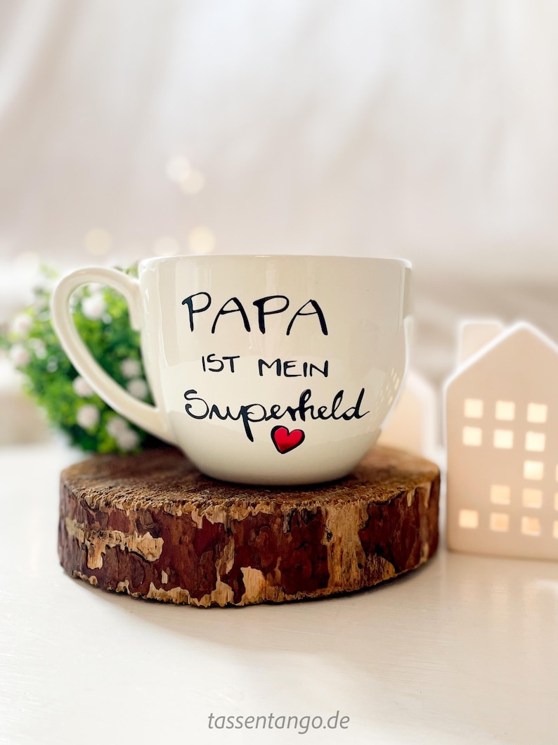 Op de afbeelding: Witte keramische mok met een zwarte inscriptie die "Papa ist mein Superheld" met een rood hart zegt. De mok staat op een houten onderzetter.