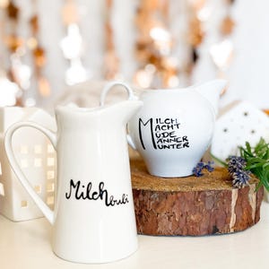 Könnte beinhalten: Zwei weiße Milchkrüge aus Keramik mit schwarzer Beschriftung. Ein Krug sagt "Milch macht ude anner unter" und der andere sagt "Milchtrubi". Die Krüge stehen auf einer Holzscheibe.