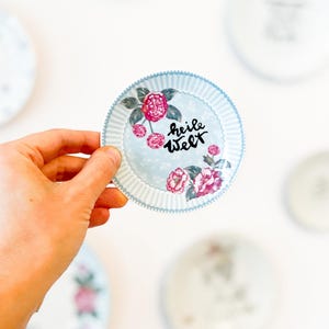 Könnte beinhalten: Ein kleiner runder Keramikteller mit einem hellblau-weiß gestreiften Rand und einem Blumenmuster aus rosa Blüten und Kirschen. In der Mitte des Tellers befindet sich der schwarze handgeschriebene Text "heile Welt" auf hellblauem Grund.