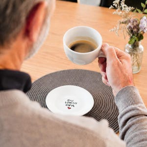May include: A white ceramic coffee cup filled with dark coffee is held by a person. A white saucer with the text "DU WIRST OPA" and a heart is on a gray placemat. A small vase with flowers is on the table.
