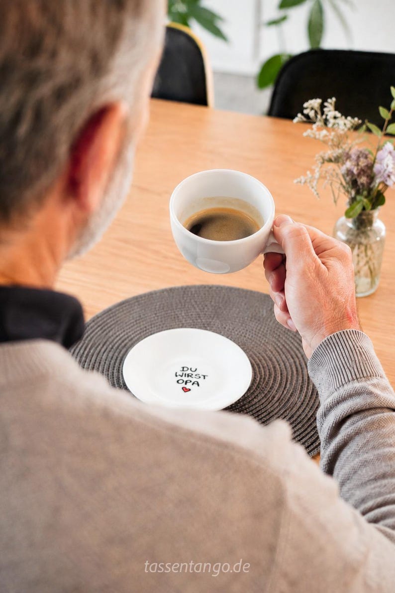 May include: A white ceramic coffee cup filled with dark coffee is held by a person. A white saucer with the text "DU WIRST OPA" and a heart is on a gray placemat. A small vase with flowers is on the table.