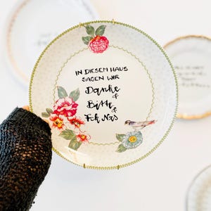Könnte beinhalten: Ein weißer Keramikteller mit grünem Wellenrand, verziert mit bunten Blumenillustrationen und einem kleinen Vogel. Der Text lautet: IN DIESEM HAUS SAGEN WIR Danke & Bitte & Fck NzS. Der Teller hat ein dezentes, gepunktetes Muster.