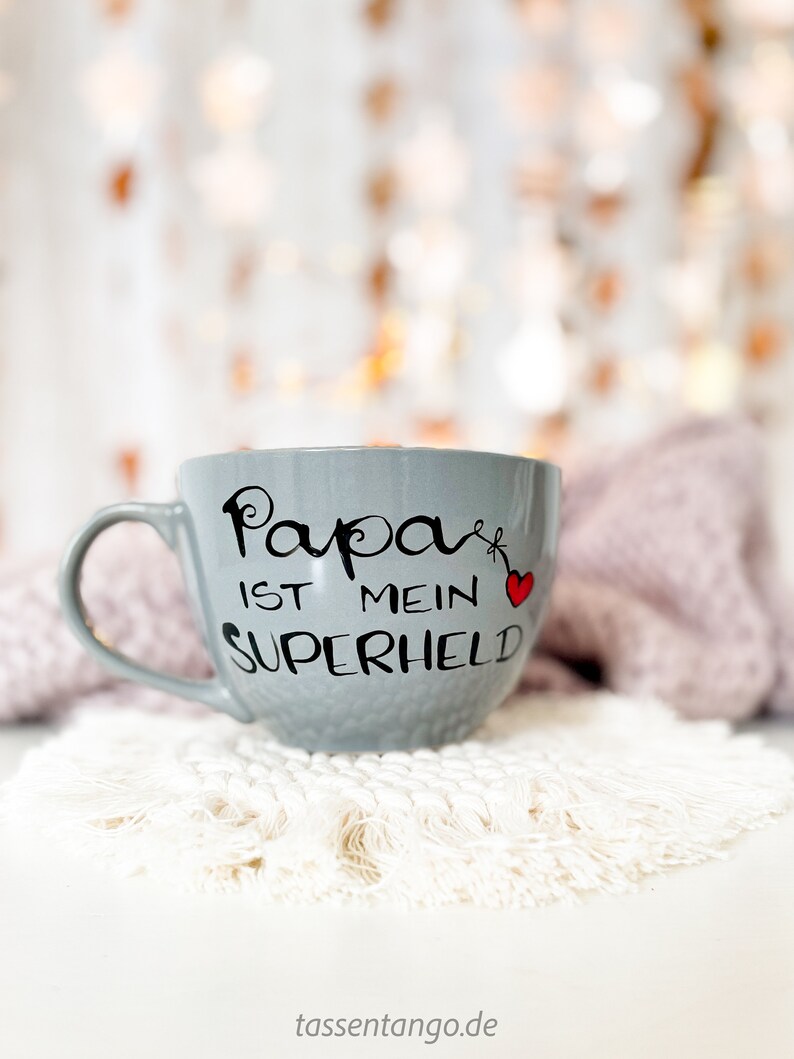 Op de afbeelding: Een grijze keramische mok met een zwart-wit ontwerp met de tekst "Papa ist mein Superheld" met een rood hart. De mok staat op een witte gehaakte onderzetter.