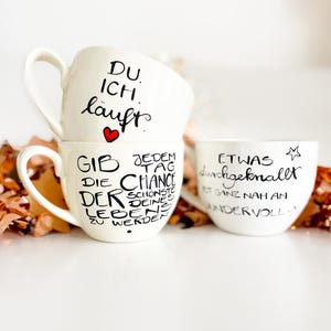 Könnte beinhalten: Drei weiße Keramikbecher mit schwarzem handgeschriebenen Text. Der obere Becher sagt "DU. ICH. läuft" mit einem roten Herzen. Der mittlere Becher sagt "GIB DIE CHANCE DER SCHÖNSTE DEINES LEBENS ZU WERDEN" und der untere Becher sagt "ETWAS durchgeknallt IST GANZ NAH AM WUNDERVOLL".