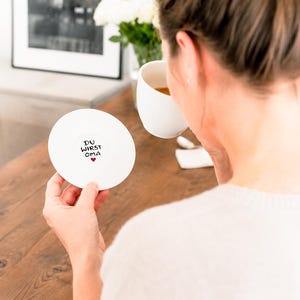 May include: A white ceramic coaster with the text "DU WIRST OMA" and a small red heart. The coaster is held by a person, with a white mug and flowers in the background. The coaster is round.