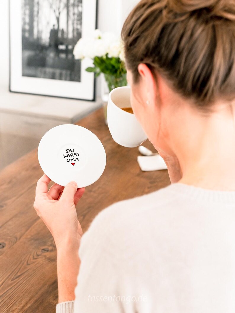 May include: A white ceramic coaster with the text "DU WIRST OMA" and a small red heart. The coaster is held by a person, with a white mug and flowers in the background. The coaster is round.