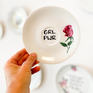 Könnte beinhalten: Eine weiße Untertasse aus Keramik mit dem schwarzen Schriftzug GRL PWR in der Mitte. Rechts neben dem Text befindet sich eine detaillierte Abbildung einer einzelnen rosa Rose mit grünen Blättern und Stiel. Die Untertasse hat eine glatte Oberfläche.