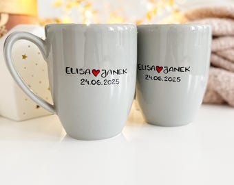 Könnte beinhalten: Zwei graue Keramikbecher mit schwarzer Schrift und einem roten Herzen. Die Becher sagen "ELISA ♥ JANEK 24.06.2025".