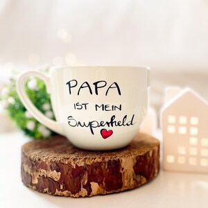 Op de afbeelding: Witte keramische mok met zwarte tekst die "Papa ist mein Superheld" met een rood hartje zegt. De mok staat op een houten onderzetter.