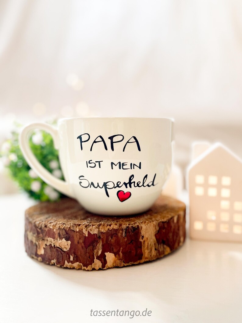 Op de afbeelding: Witte keramische mok met zwarte tekst die "Papa ist mein Superheld" met een rood hartje zegt. De mok staat op een houten onderzetter.